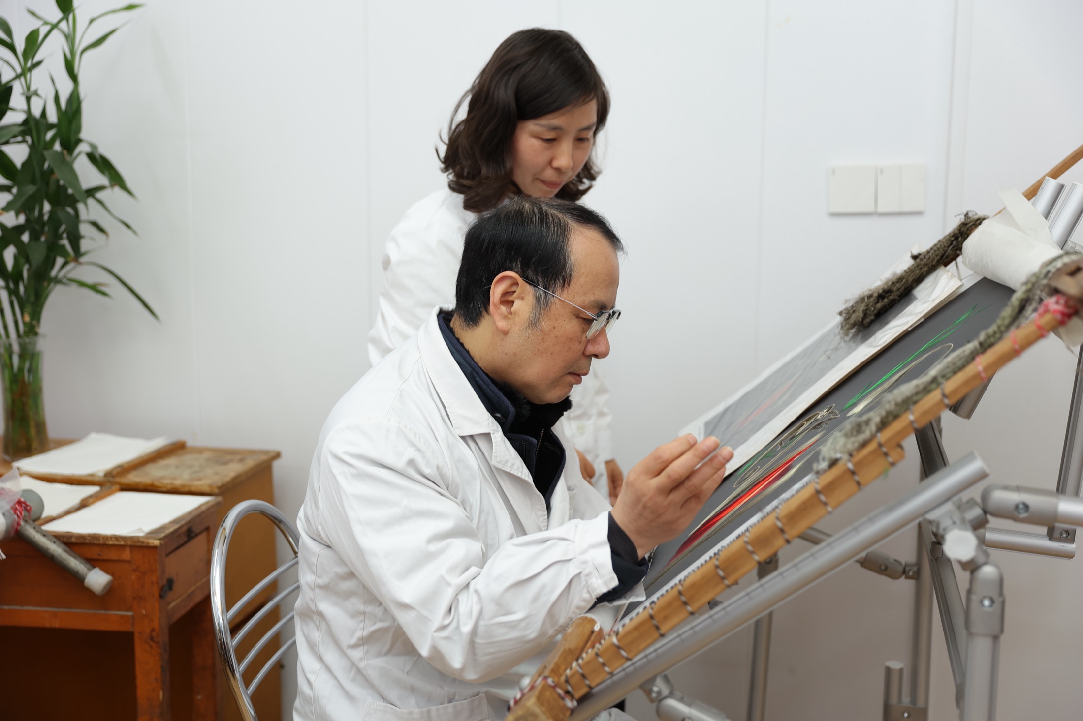 Two embroidery studio practitioners examining silk work on an angled loom.
